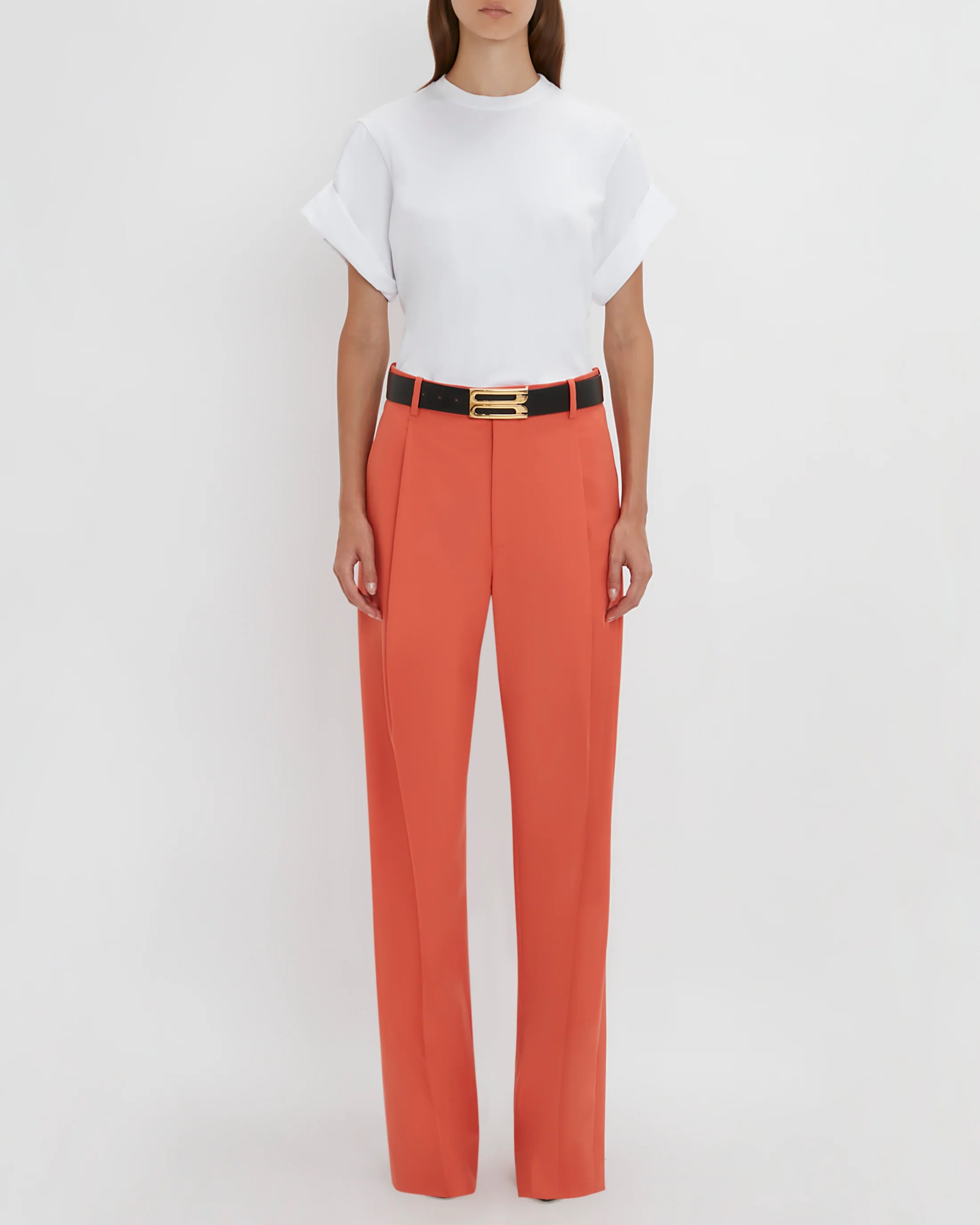 Model wears orange single pleat trouser with black belt with gold buckle and white short sleeved t-shirt. 