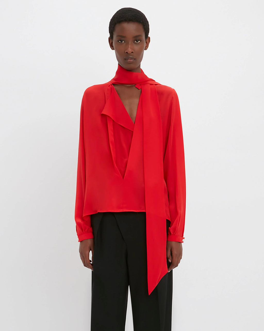 Model wears red long sleeve top with scarf neck design detail and black pants. Set against a white background. 