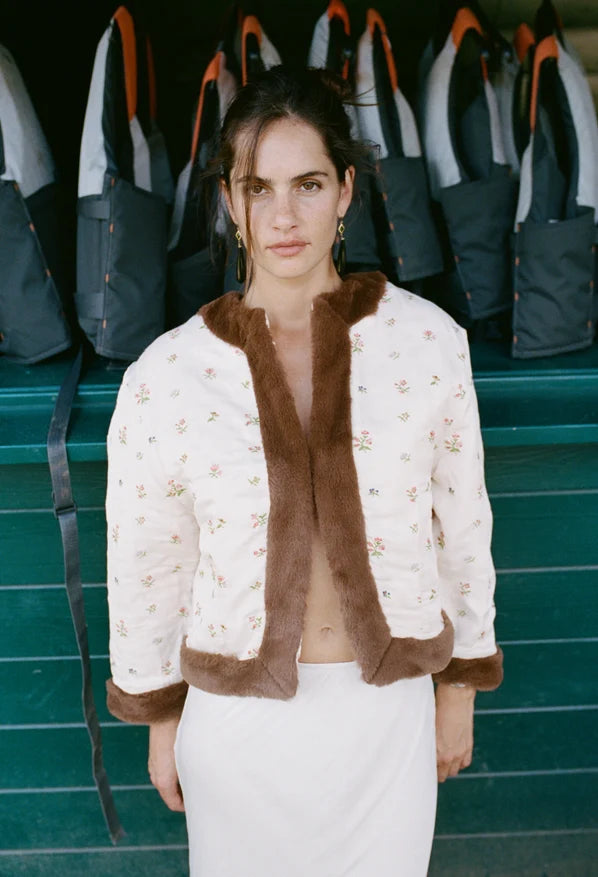 A woman stands in front of a teal bench with dark gray bags, wearing a cream quilted jacket with brown faux-fur trim and a small floral print, paired with a white skirt.