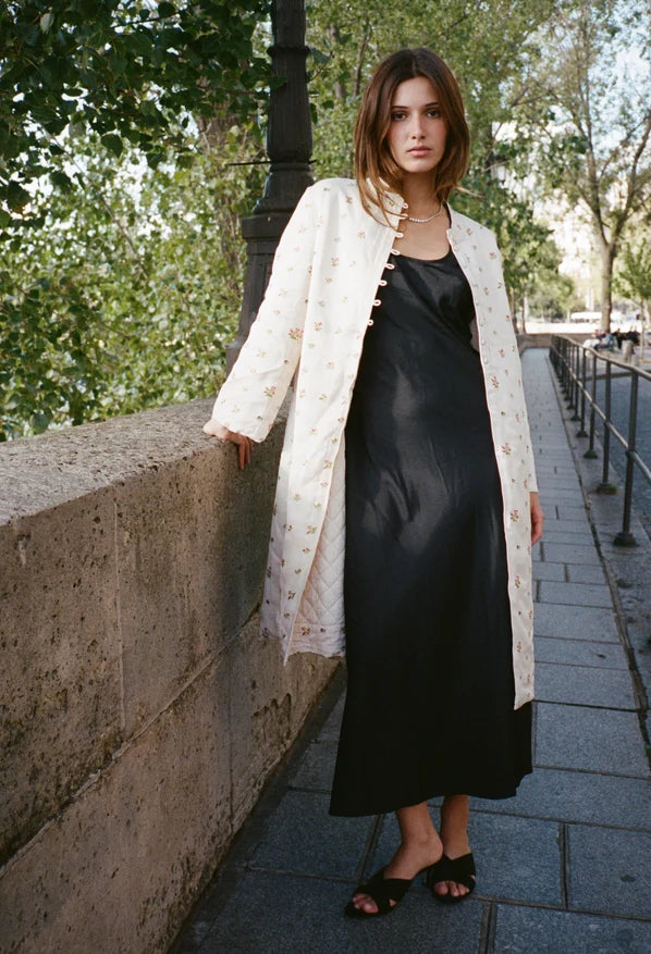 A woman stands on an outdoor stone walkway wearing a long cream embroidered jacket over a black satin slip dress and black sandals.