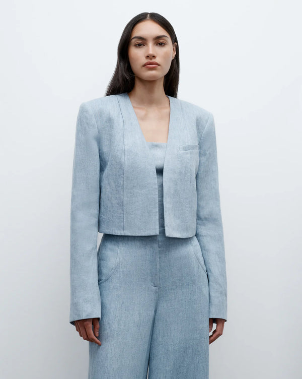PHOTO OF BROWN HAIRED MODEL ON A WHITE WALL. SHE IS WEARING THE BLUE LINEN BLAZER AND A BLUE LINEN TROUSER. THE BLAZER IS SITTING AT HER WAISTBAND. 