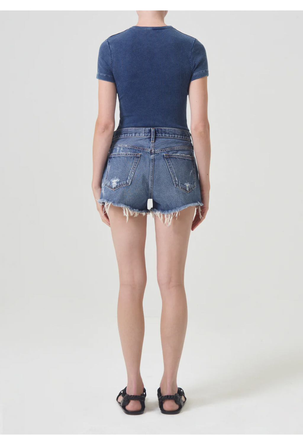 Back side of model wearing dark wash cut off jean shorts with back pockets and frayed raw cut hem and blue short sleeve t-shirt with black sandals. She stands against a white background. 