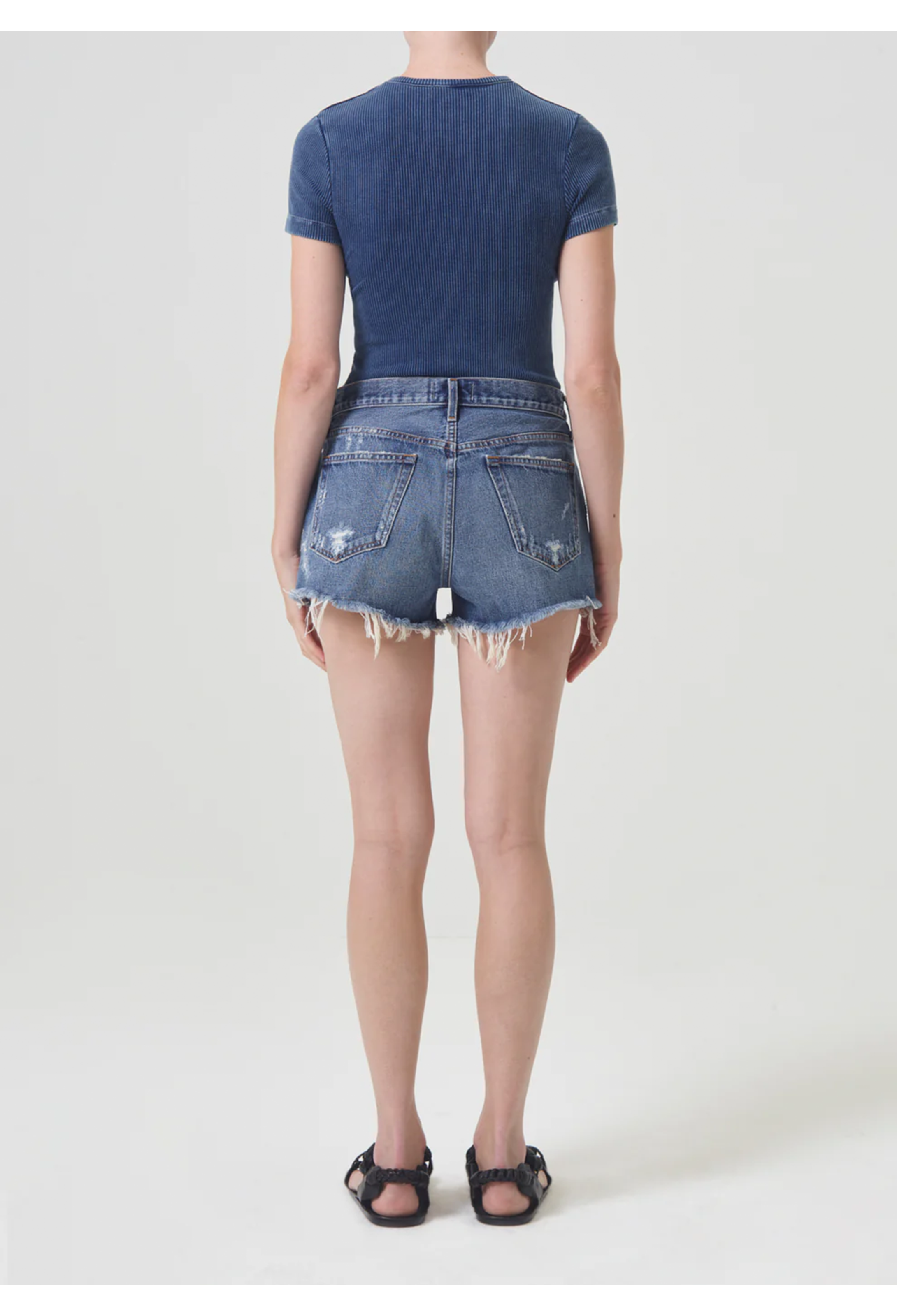 Back side of model wearing dark wash cut off jean shorts with back pockets and frayed raw cut hem and blue short sleeve t-shirt with black sandals. She stands against a white background. 