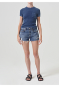 Model wears dark wash cut off jean shorts with frayed raw cut hem and blue short sleeve t-shirt with black sandals. She stands against a white background. 