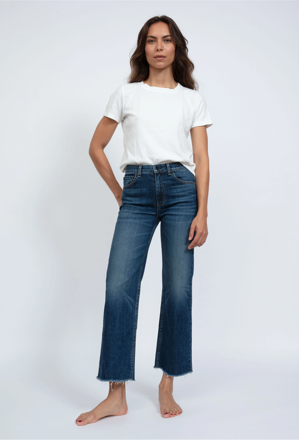 Model wears high rise wide leg cropped medium wash jeans with frayed hem, and a white short sleeve t-shirt. She stands in front of a white background. 