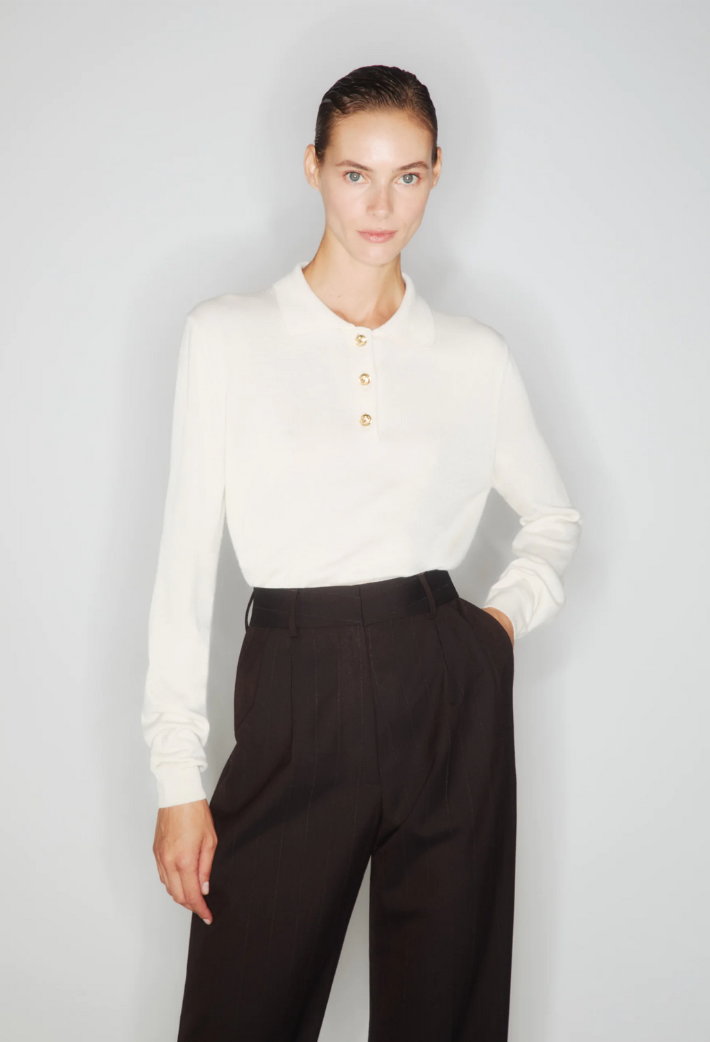 a woman wearing a white long sleeve collared button up pullover with brown pants. white background