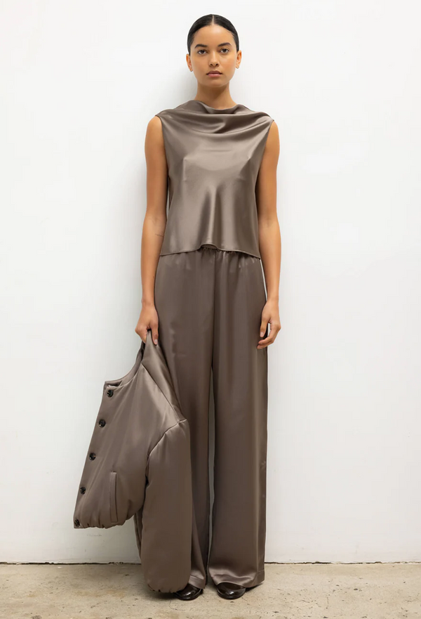 Woman wearing a brown sleeveless top and wide-leg pants holding a matching coat against a white background