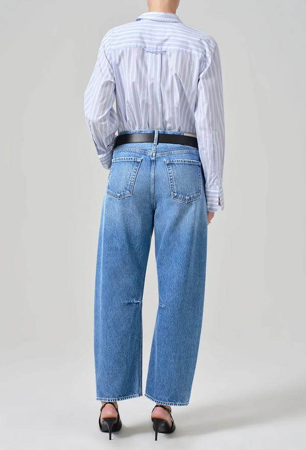 Backside of model wearing high waisted light wash wide leg barrel jeans and a black belt with a blue and white long sleeve blouse and black heels. She stands in front of a white wall. 