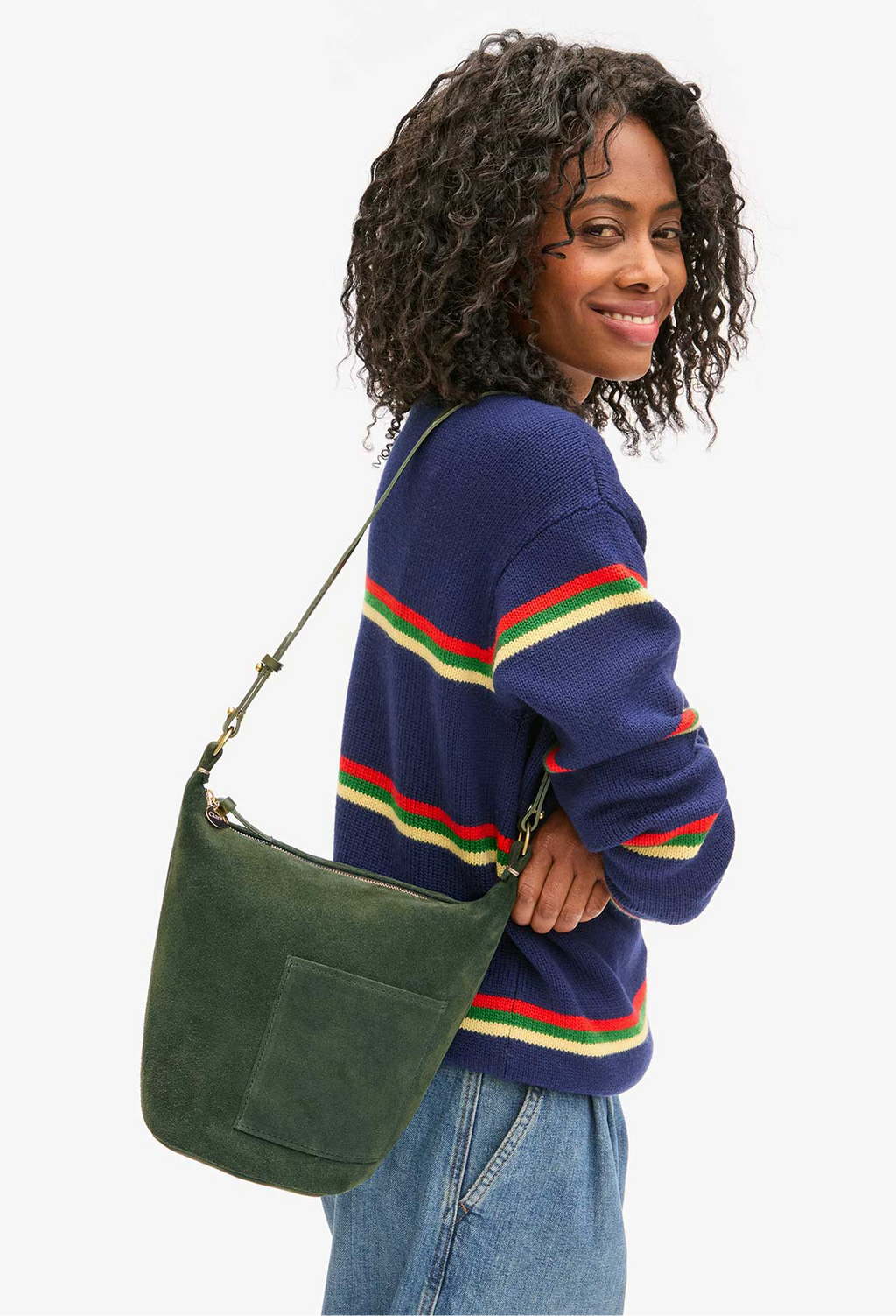 Model carries green suede bucket bag on shoulder strap. She wears a navy sweater with red, green, and yellow stripes and jeans. She stands in front of a white background. 