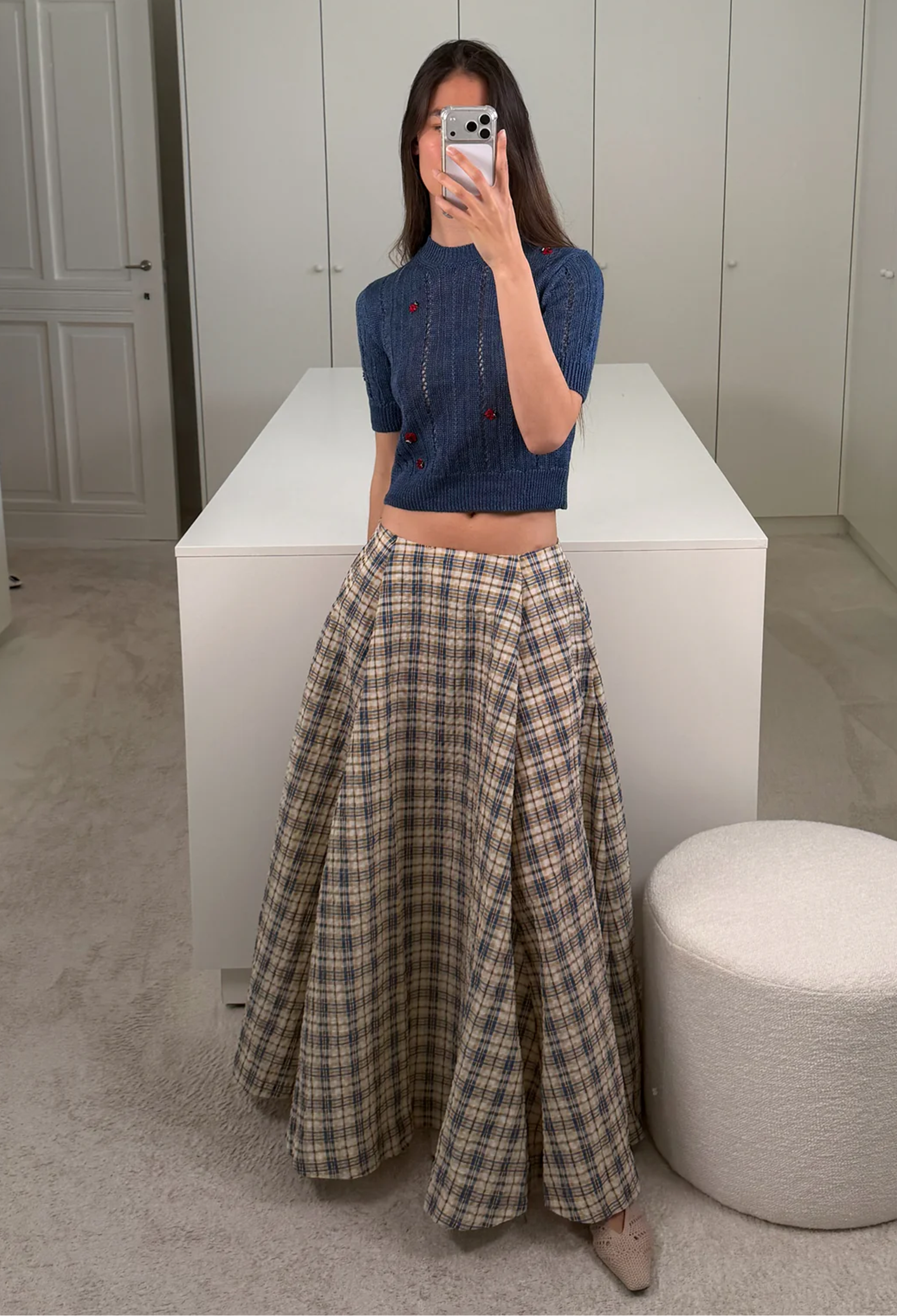 Woman taking a mirror selfie wearing a blue top and plaid skirt in a minimalistic room.