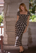 A woman models a black midi dress with a white floral print, featuring a square neckline and short puff sleeves, set against a rustic concrete pillar background.