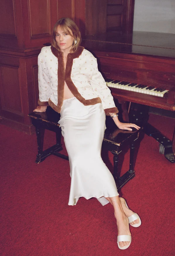 A woman sits on a piano bench in a room with dark wood paneling and red carpet, wearing a white patterned jacket with brown fur trim and a white satin skirt.