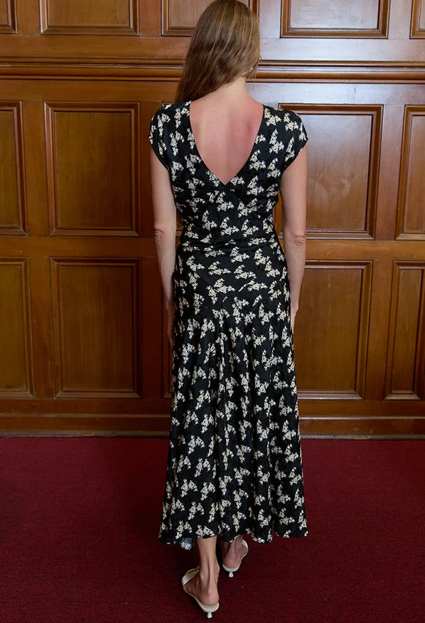 A woman viewed from behind wearing a black midi dress with a white floral pattern, standing in front of a dark wood paneled wall on a red carpet.