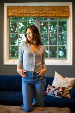 a model in front of a blue couch and a window wearing a grey v-neck cardigan and blue jeans.