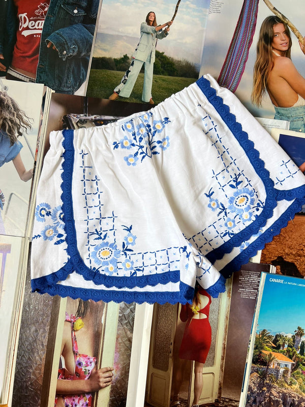 a pair of white stretch waist shorts with light blue floral embroidery and dark blue border hemming laying on top of fashion magazines.