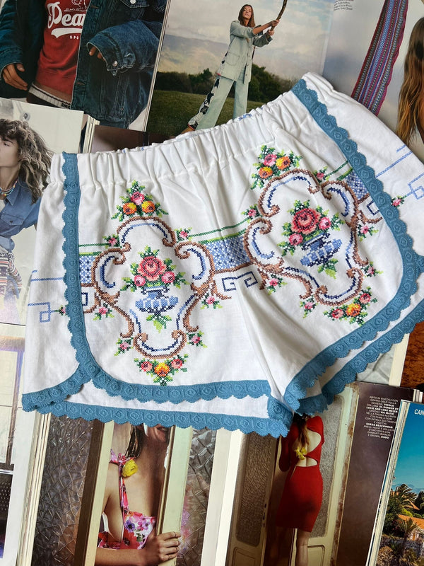 a pair of white stretch waist shorts with multicolored floral embroidery and blue border hemming laying on top of fashion magazines.