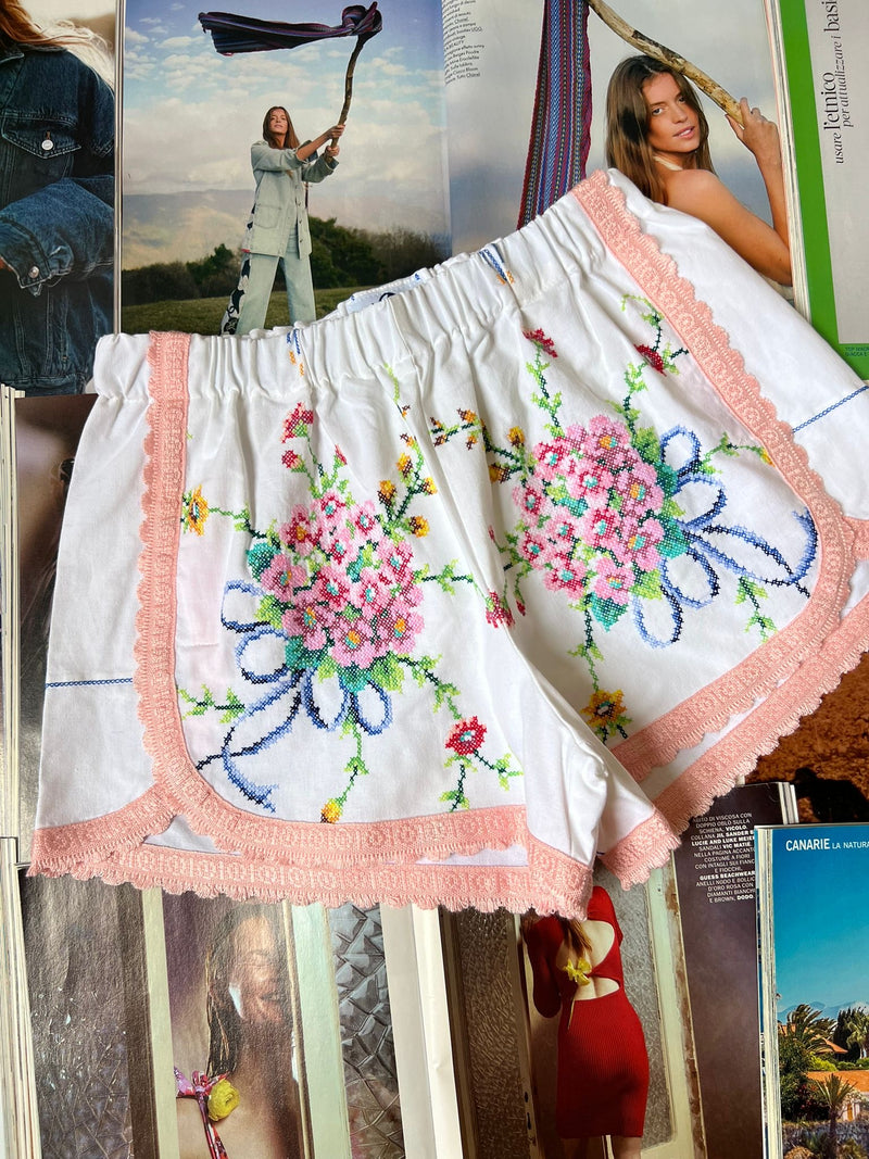a pair of white stretch waist shorts with multicolored floral embroidery and light pink border hemming laying on top of fashion magazines.
