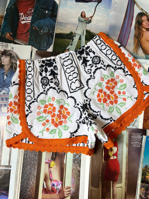 a pair of white stretch waist shorts with black geometric and orange floral embroidery and orange border hemming laying on top of fashion magazines.