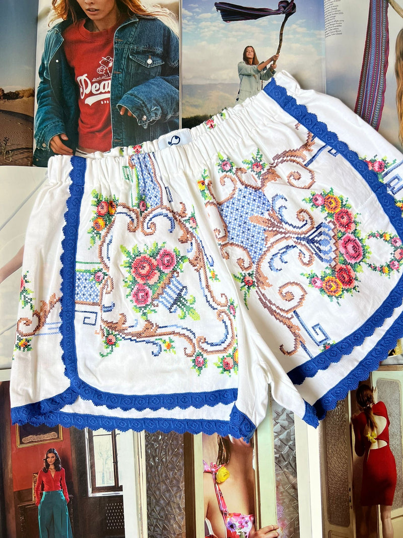 a pair of white stretch waist shorts with multicolored floral embroidery and navy border hemming laying on top of fashion magazines.