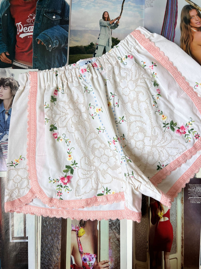 a pair of white stretch waist shorts with multicolored floral embroidery and light pink border hemming laying on top of fashion magazines.