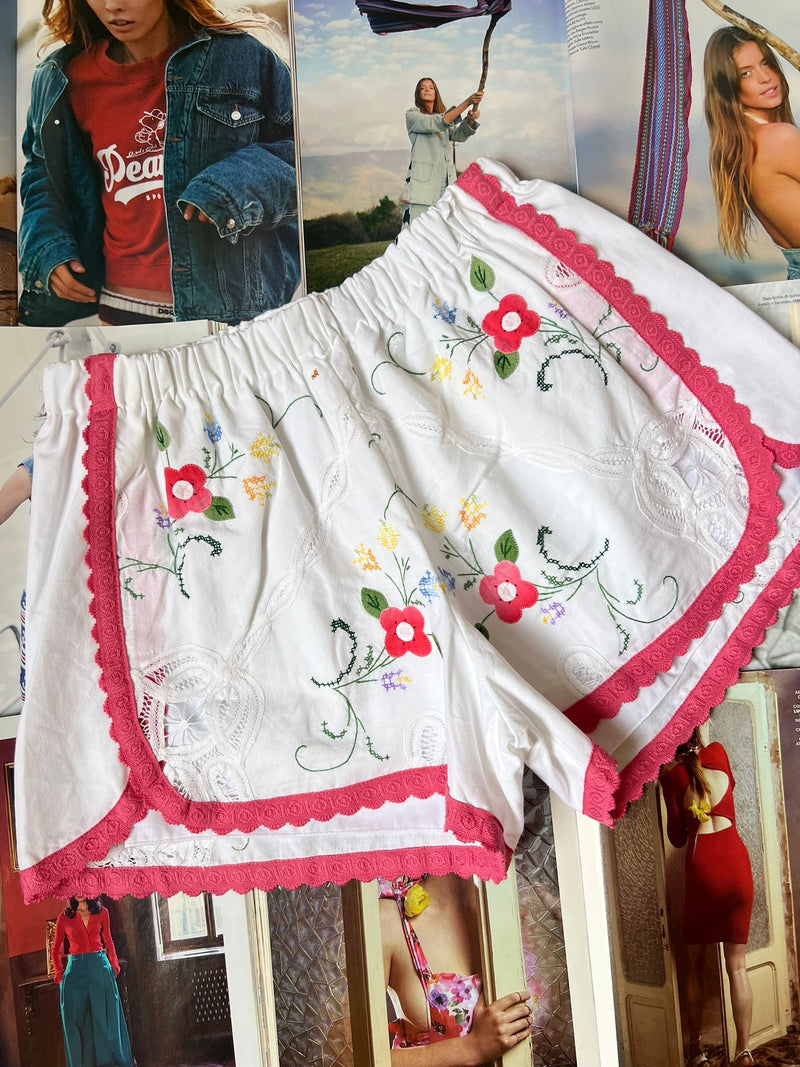 a pair of white stretch waist shorts with multicolored floral embroidery and hot pink border hemming laying on top of fashion magazines.