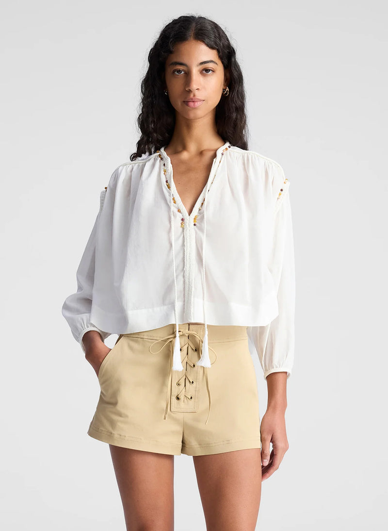 MODEL WEARING A 3/4 SLEEVE WHITE TOP WITH BEADED EMBELLISHMENTS ON THE NECKLINE AND SHOULD LINE. THE SHIRT HAS TASSELS THAT ARE UNTIED AND MODEL HAS ON TAUPE TIE UP SHIRT SHORTS. SHE IS AGAINST A LIGHT GREY BACKGROUND. 