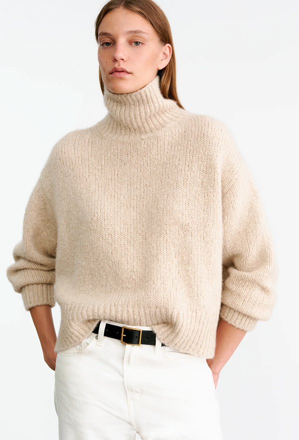 Person wearing a beige turtleneck sweater and white pants on a plain background