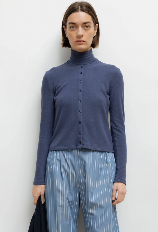 Person wearing a blue cardigan over a turtleneck and striped pants against a white background