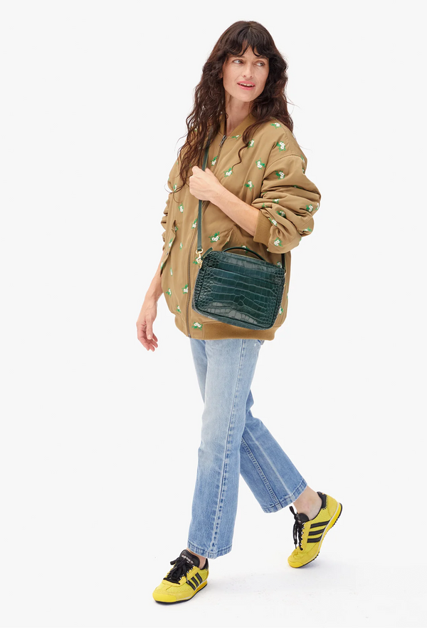 Woman holding a green crossbody bag wearing a patterned sweater and jeans on a white background
