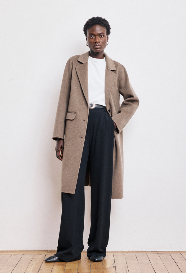 Person wearing a beige coat, white shirt, and black pants against a white wall.