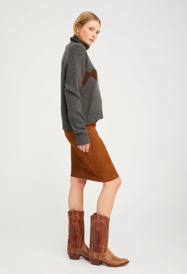 Woman wearing a gray sweater with a red logo and a brown skirt, standing against a white background.