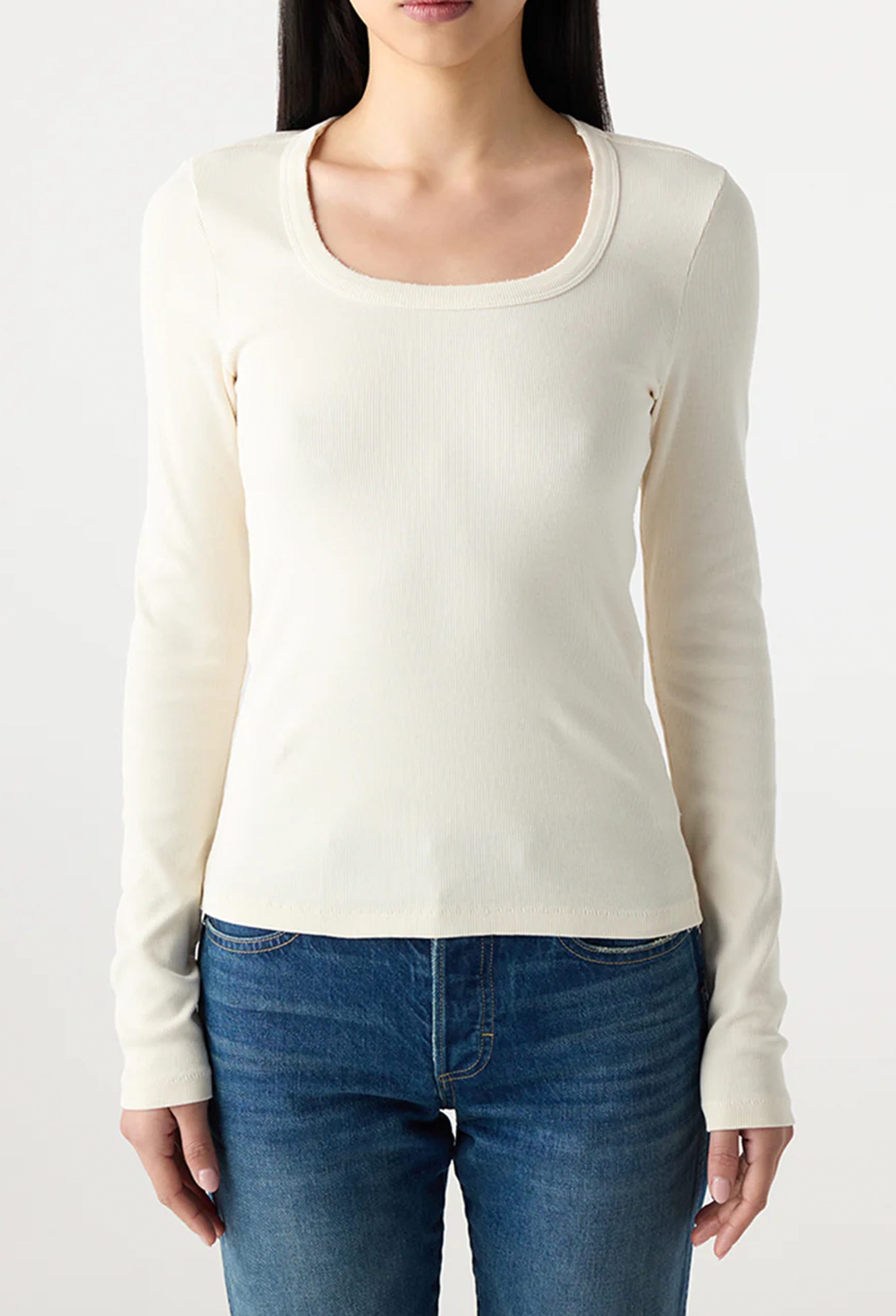 Woman wearing a white long-sleeve top and blue jeans on a plain background