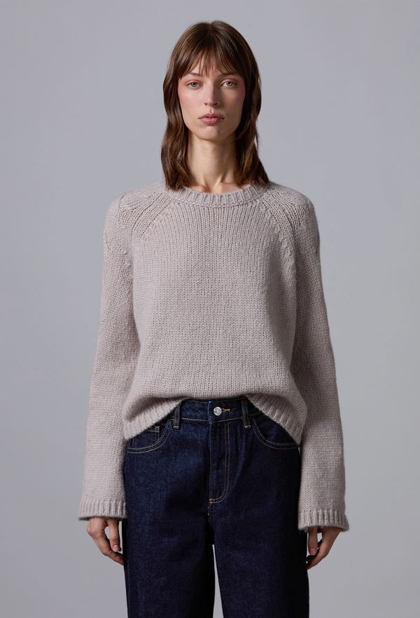 Woman wearing a light beige sweater and dark blue jeans against a plain background