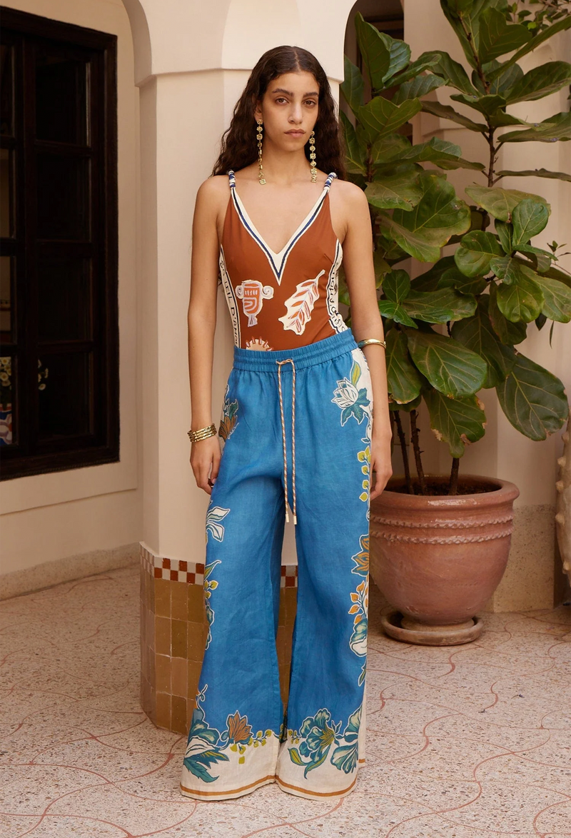 Woman wearing a vintage-style outfit with a brown top and blue pants with floral embroidery, standing in a decorative outdoor setting.