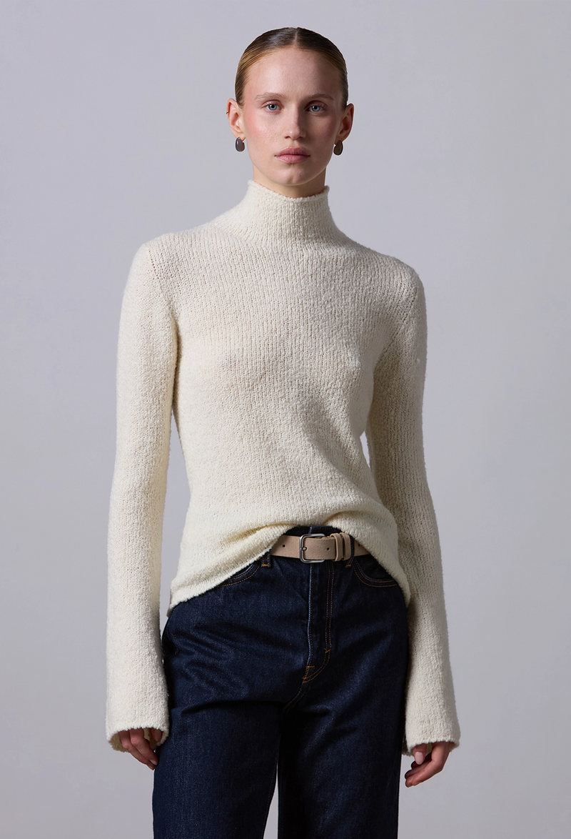 Person wearing a cream turtleneck sweater and dark pants against a plain background