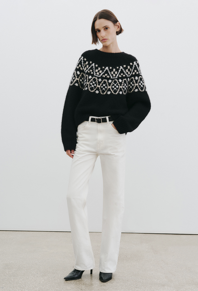Person wearing a black sweater with fair isle pattern and white pants on a plain background