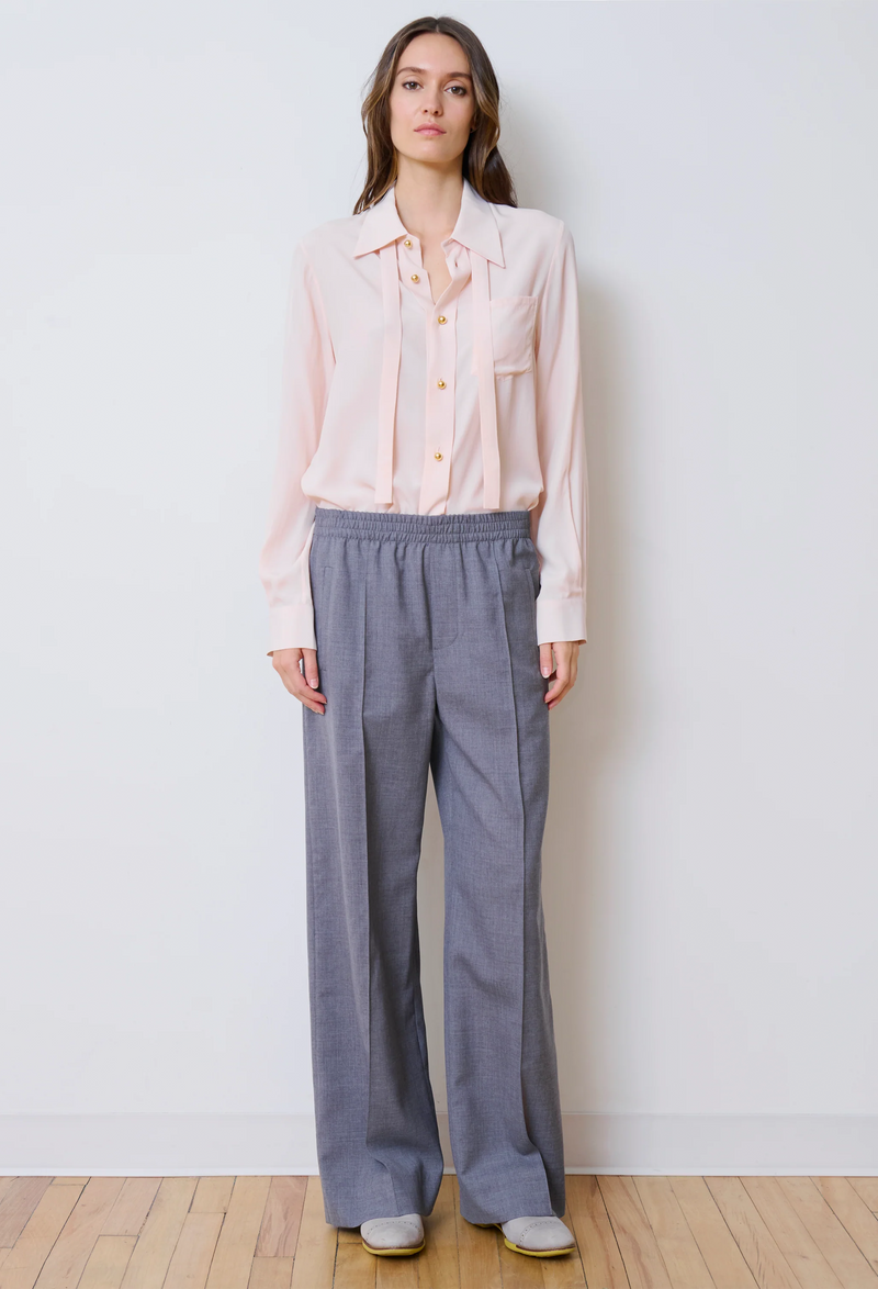 Woman wearing a light pink blouse and gray pants against a white wall.