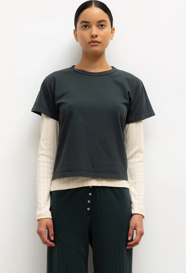 Person wearing a green t-shirt over a cream long-sleeve shirt with dark green pants on a white background