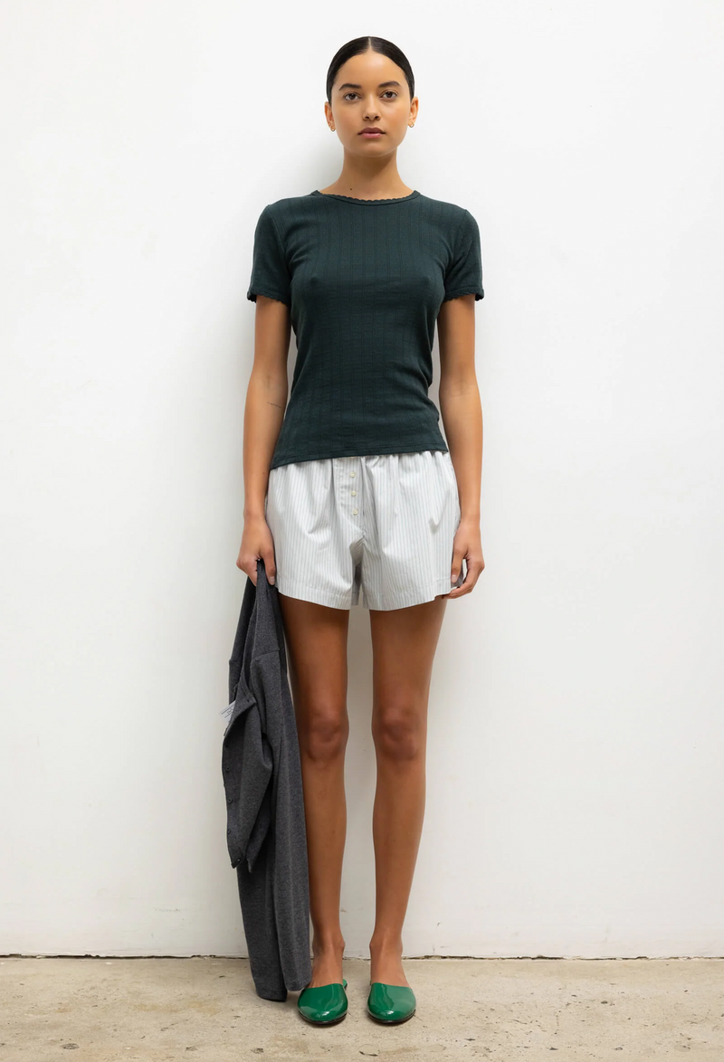 Woman wearing a dark green t-shirt and white shorts, standing against a plain background.