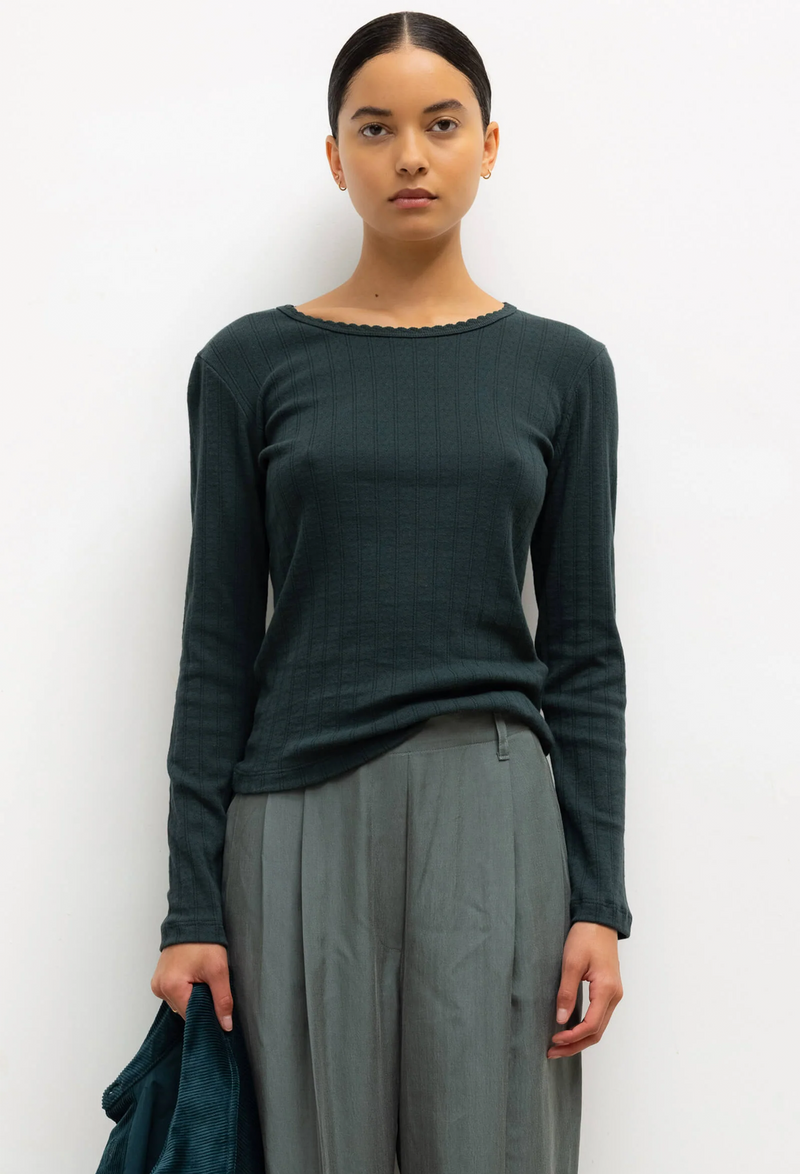 Woman wearing a dark green long-sleeve top and gray pants against a white background