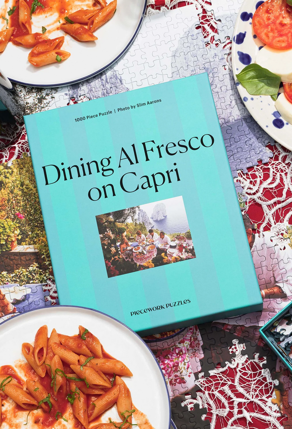 Jigsaw puzzle box titled 'Dining Al Fresco on Capri' surrounded by plates of pasta.