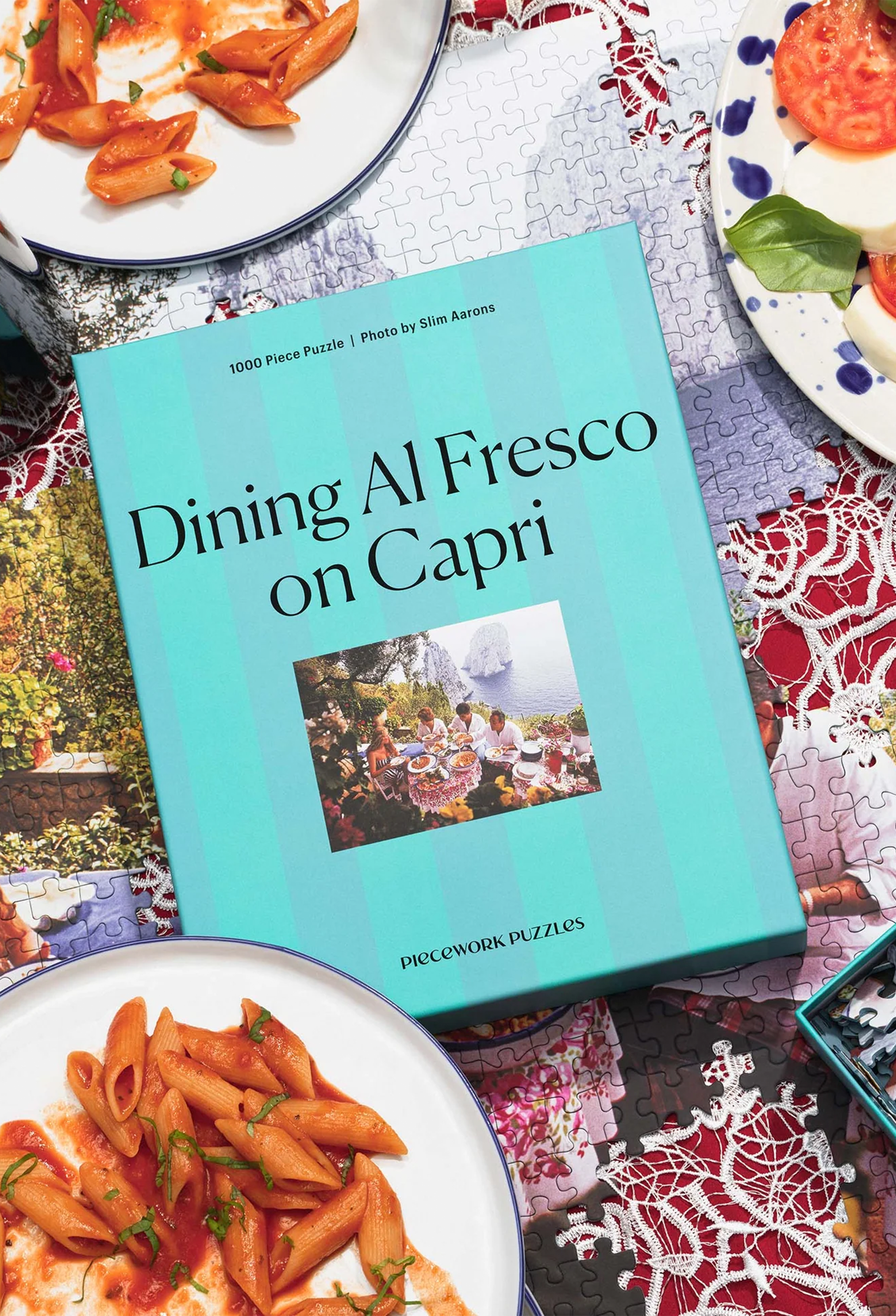 Jigsaw puzzle box titled 'Dining Al Fresco on Capri' surrounded by plates of pasta.