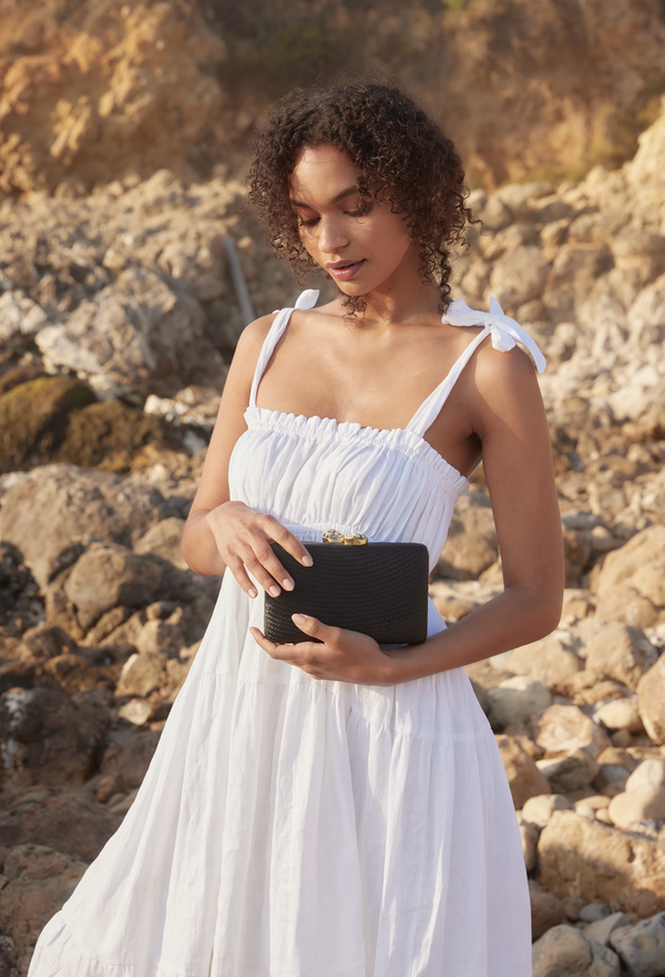 Woman in a white dress holding a black clutch against a rocky background