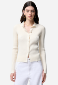 Woman wearing a cream-colored ribbed cardigan over a white shirt with white pants on a light gray background