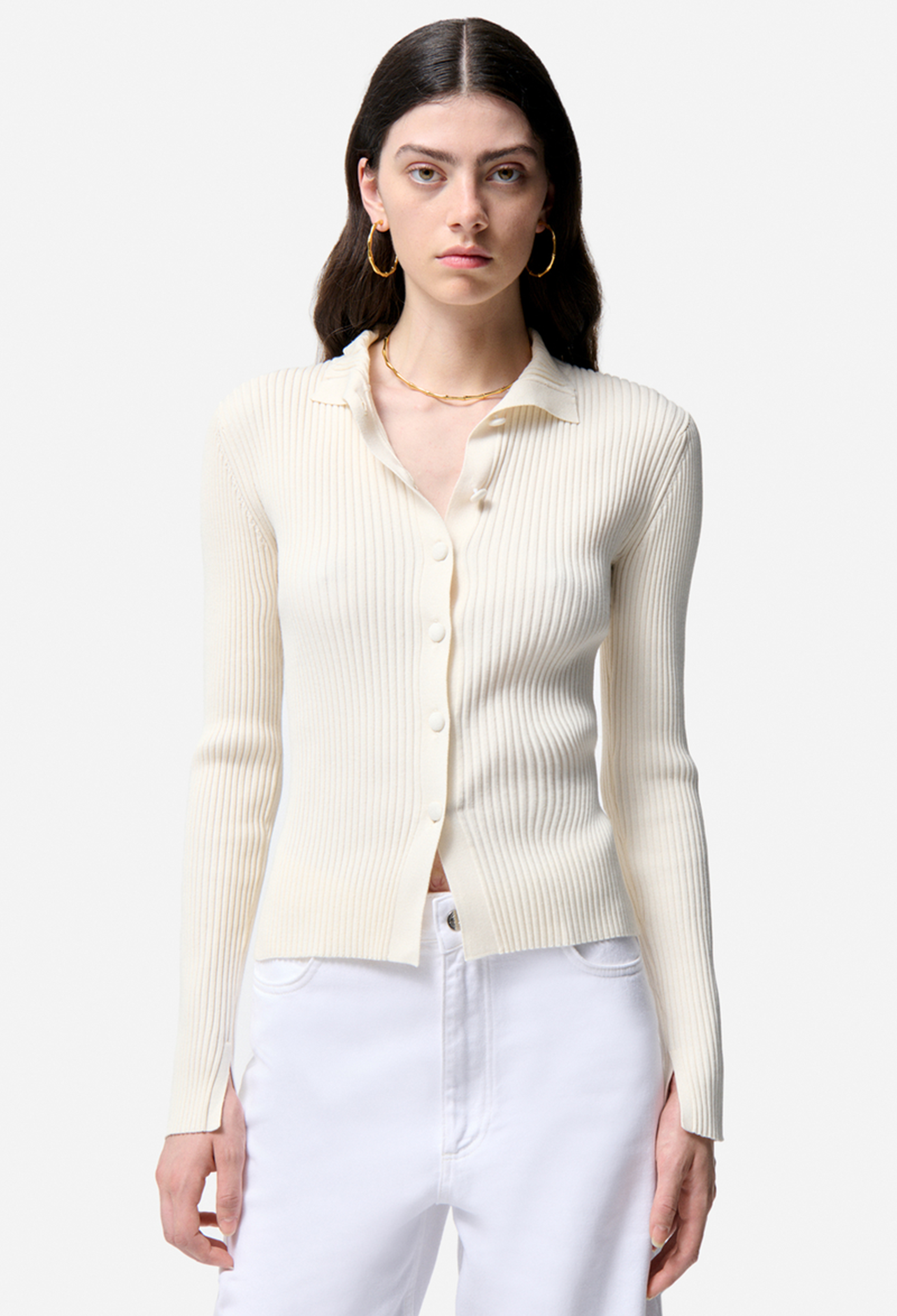 Woman wearing a cream-colored ribbed cardigan over a white shirt with white pants on a light gray background