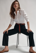 Woman wearing a white shirt and black pants with a brown belt, sitting on a chair against a plain background.