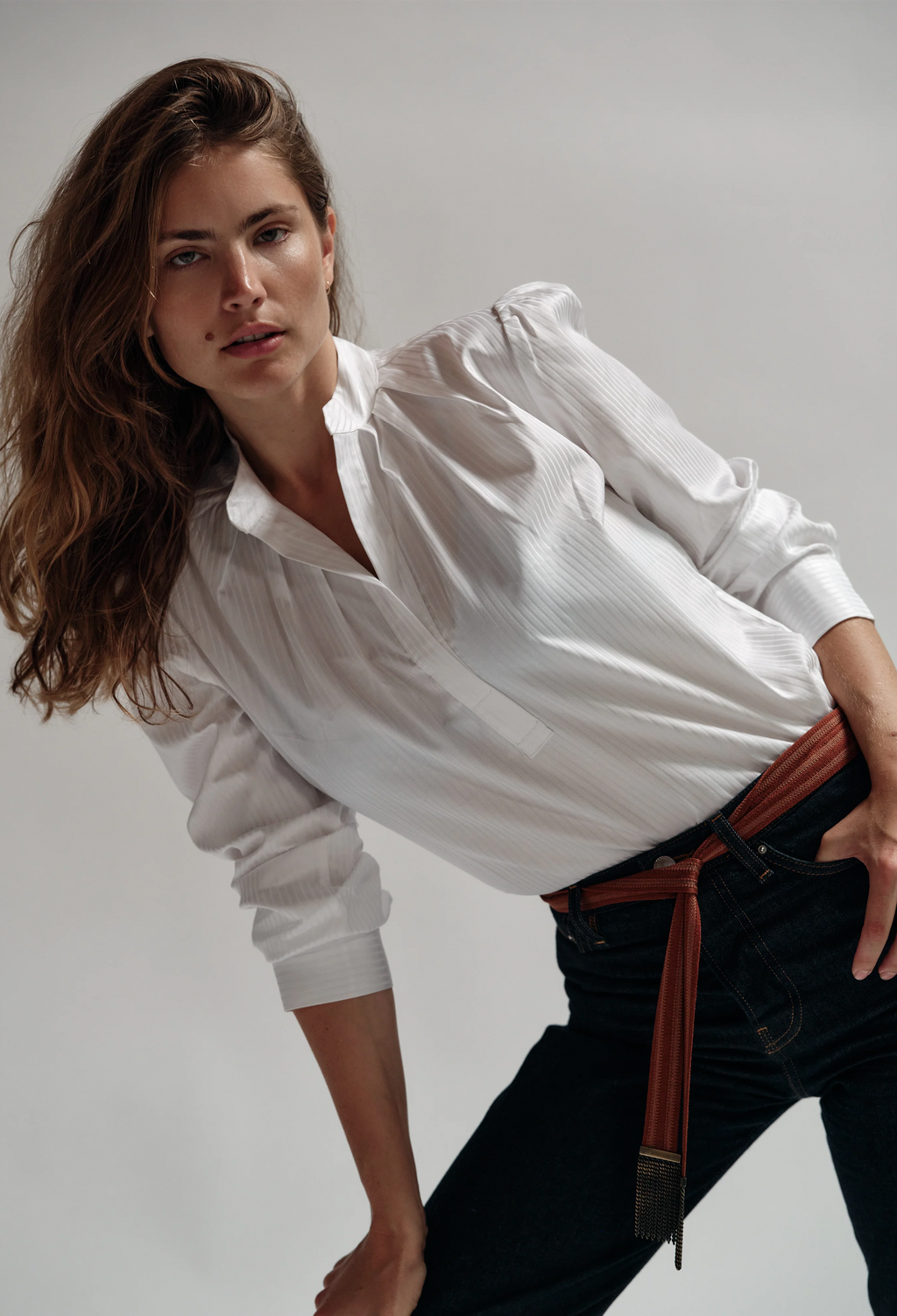 Woman wearing a white blouse and black pants with a brown belt against a plain background