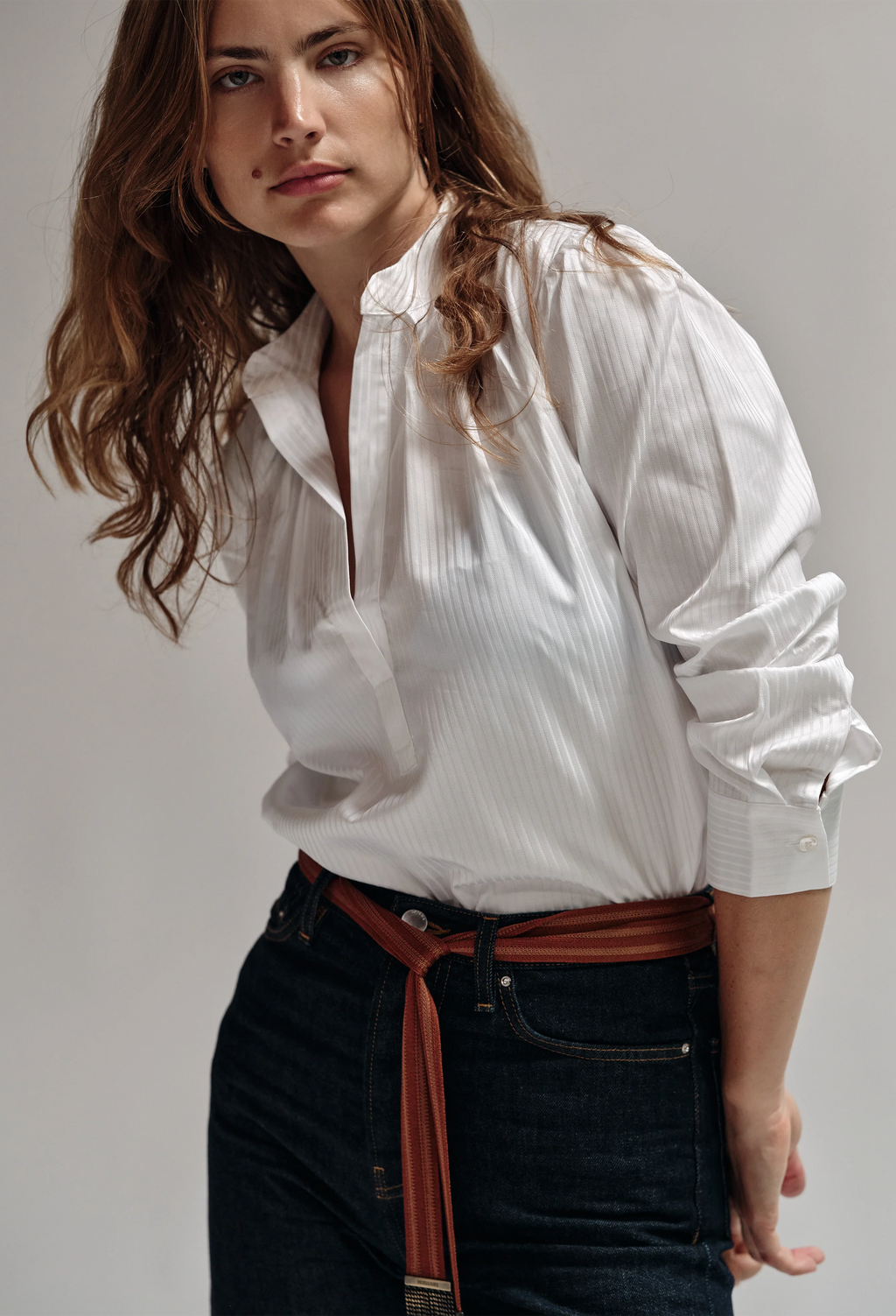 Woman wearing a white shirt and dark jeans with a brown belt against a plain background