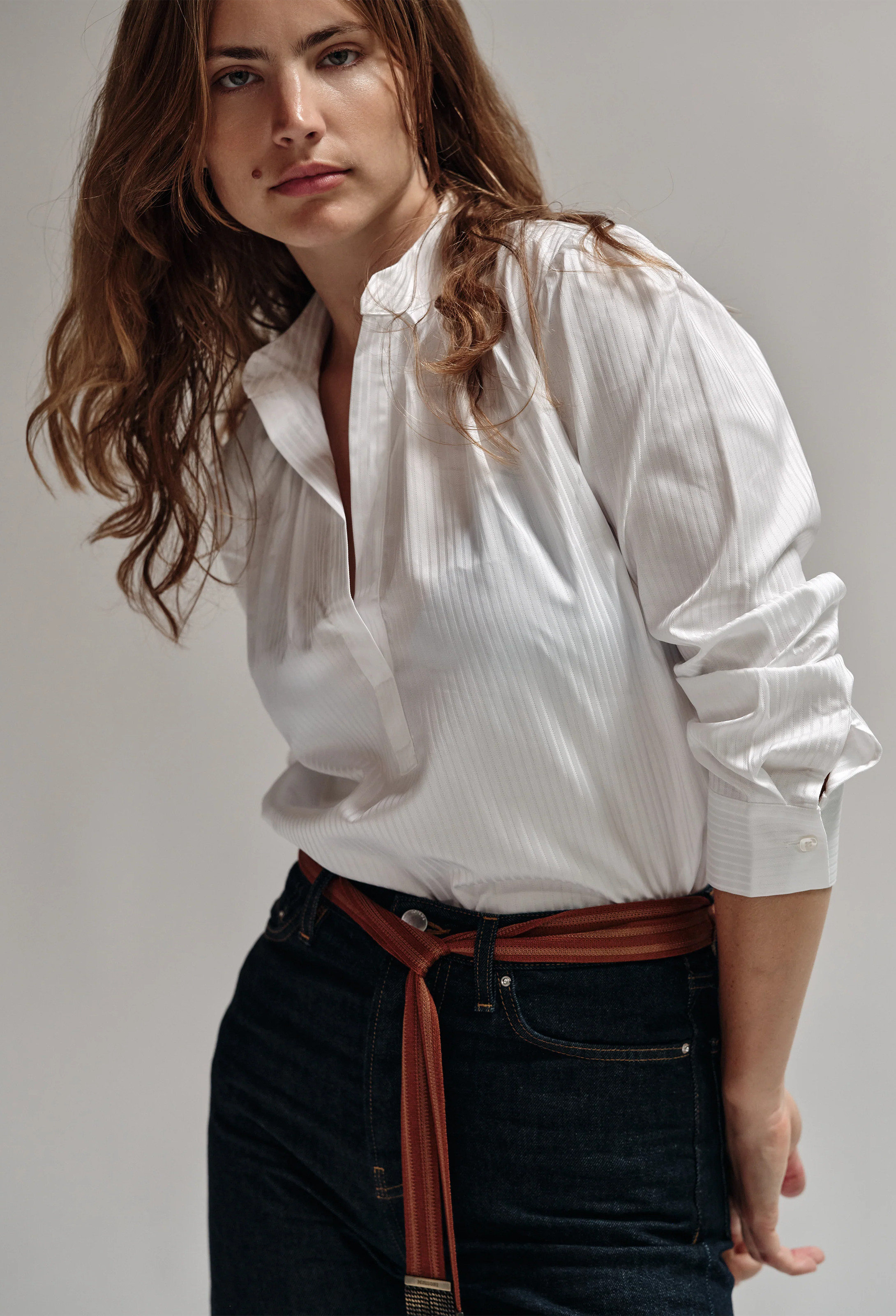 Woman wearing a white shirt and dark jeans with a brown belt against a plain background