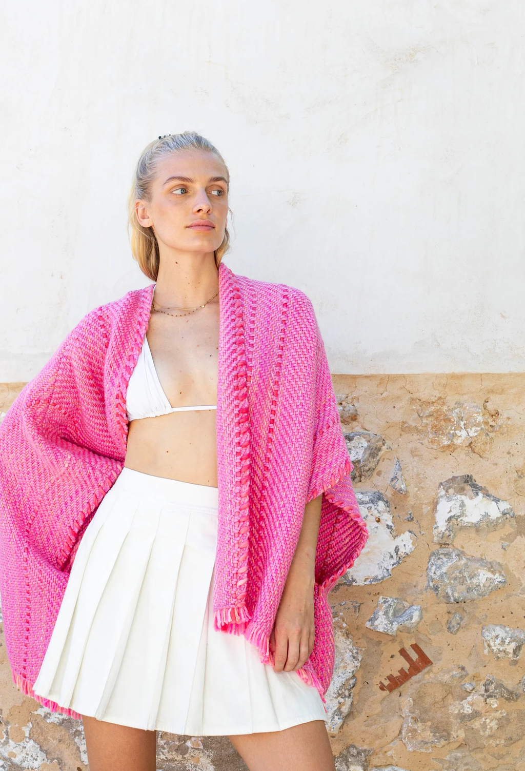 Woman wearing a bright pink knitted shawl over a white outfit against a textured wall.